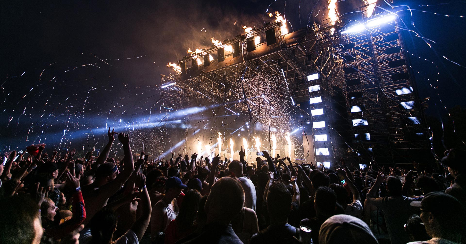 A large crowd at a nighttime outdoor concert with fireworks and a light show