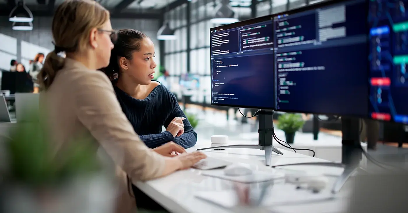 Two engineers collaborate at a workstation, reviewing AI-assisted code across multiple screens.