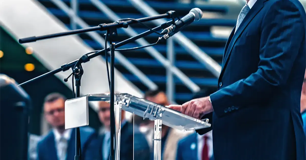 A suited figure stands at a podium with microphones, symbolizing leadership under threat and the growing need for executive protection.