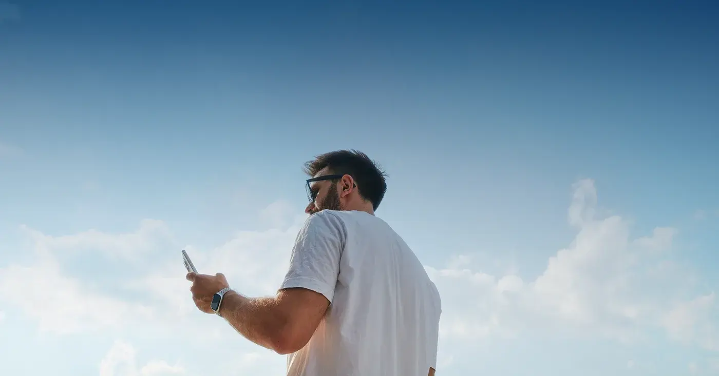 Person holding a phone under a blue sky, representing emerging social platforms like Bluesky and their role in threat monitoring.