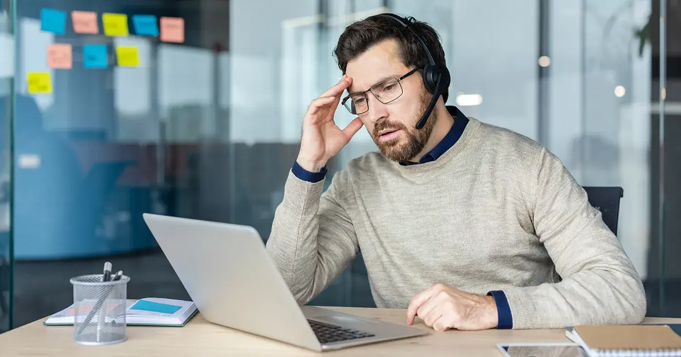 An employee wearing a headset appears distressed while working on a laptop in an office.