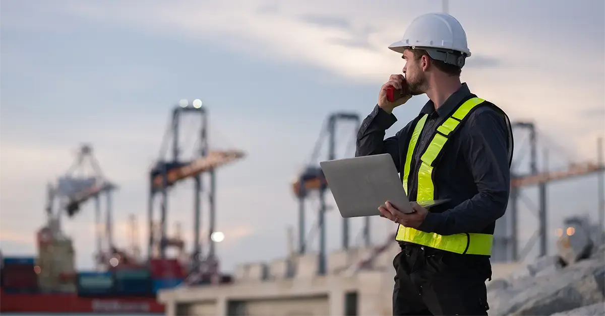 An engineer standing at an industrial port holding a laptop and radio, monitoring operations, representing IT and OT convergence and the need for unified security visibility.