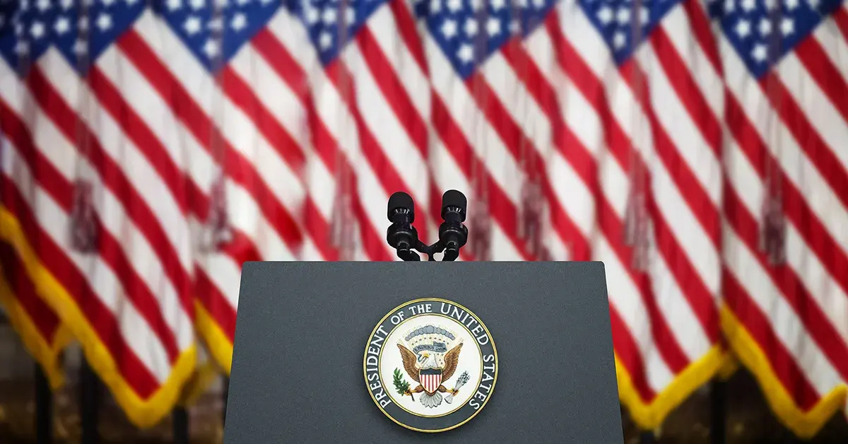 Podium with the Seal of the President of the United States and microphones in front of multiple American flags, representing the setting of a State of the Union address.